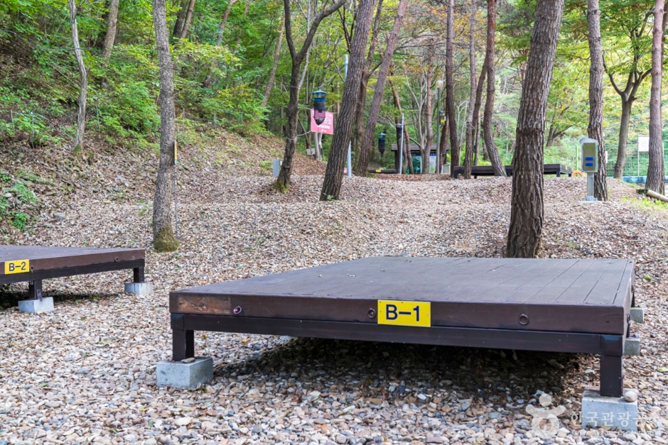 숲 경사지의 자갈 바닥 위에 낮게 놓인 B-1 야영데크와 뒤쪽 B구역